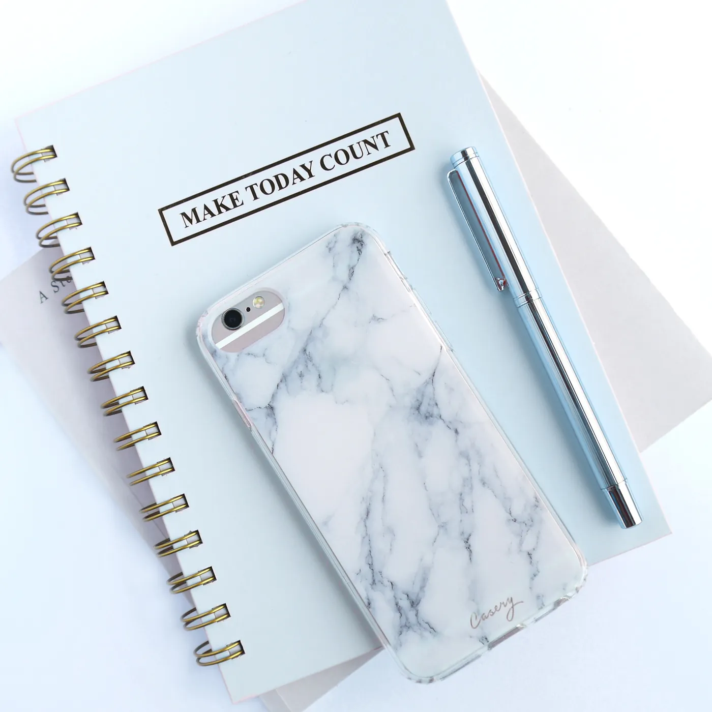 A pink notebook reading Make Today Count, a phone in a marble case and a rose-gold pen